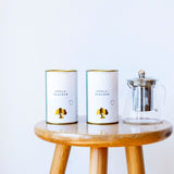 Two white cylindrical cans labeled 'IMPALA PEACOCK' with gold accents sit on a round wooden stool next to a clear glass teapot with a metal infuser, against a plain white background.