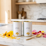 Two white canisters of Impala Peacock organic tea with gold lids, standing on a marble countertop with yellow flowers, rose petals, and a gold spoon in a warm, blurred kitchen setting.