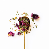 A gold spoon filled with loose-leaf rose tea, with dried purple rose petals and green leaves scattered around it on a clean white background.