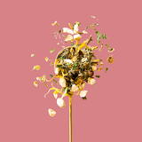 A gold spoon filled with loose-leaf herbal tea or spices, with pieces exploding outward in a dynamic splash, set against a solid dusty pink background.