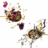 Two gold spoons filled with loose-leaf herbal tea, with dried rose petals and tea leaves splashing out against a solid white background.
