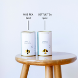 Two white cylindrical tea cans labeled 'RISE TEA (am)' and 'SETTLE TEA (pm)' sit on a round wooden stool against a light gray background. The cans are branded Impala Peacock Tea.